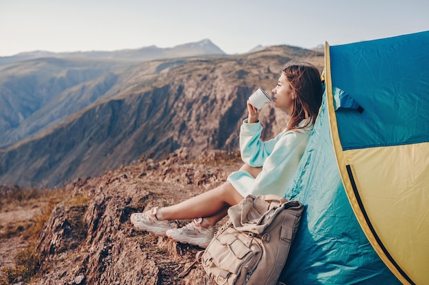 A tourist female dressed in a blue oversize hoodie is sitting near a tourist tent and drinking hot tea from a mug while admiring the mountain landscape.