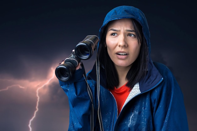 A tourist girl in a blue raincoat holds binoculars in her hands and looks into the distance spies