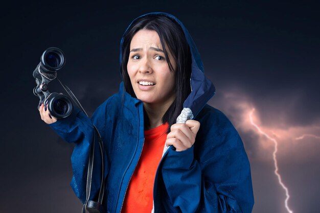 A tourist girl in a blue raincoat holds binoculars in her hands and looks into the distance spies