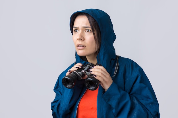 A tourist girl in a blue raincoat holds binoculars in her hands and looks into the distance spies