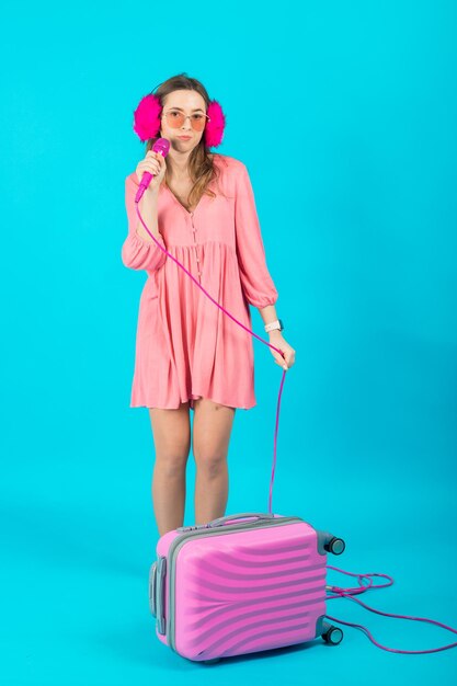 Tourist girl in a pink dress with a pink microphone on a blue background holding a pink suitcase