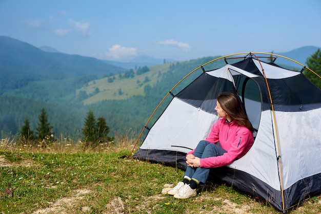 Tourist girl sitting in the entrance of small tent and enjoying beautiful foggy mountains covered with forest view under clear blue sky on bright summer morning