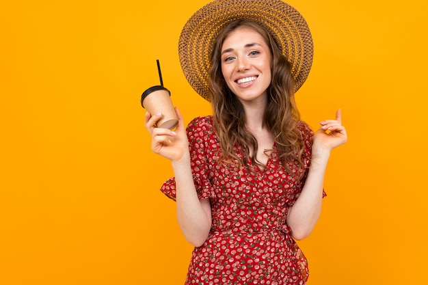 Tourist girl in a straw hat in a red dress with a glass of cooling drink on a yellow with copy space