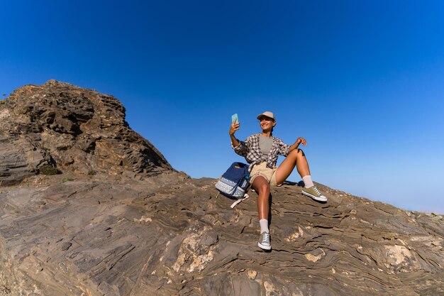 Photo tourist girl with a backpack in the summer on top of a mountain takes a selfie against the backdrop