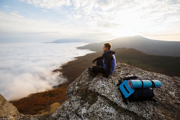 Турист с рюкзаком отдыхает на вершине горы, глядя на пейзажный закат над облаками