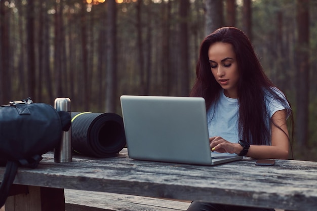 Tourist hipster girl in white shirt working on a laptop while sitting on a wooden bench in the beautiful autumn forest.