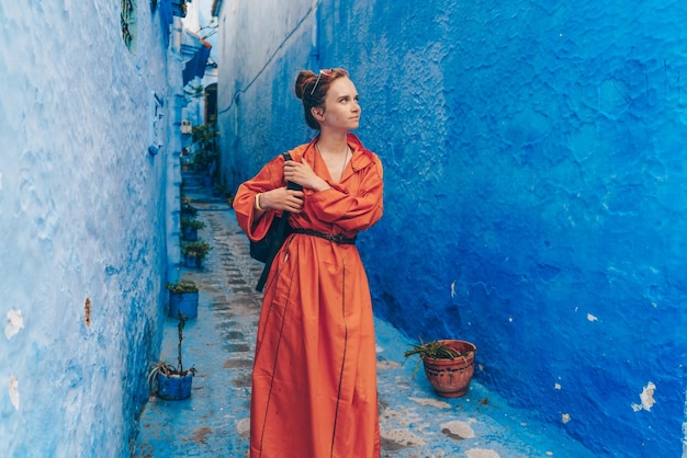 A tourist in a long bright orange dress with a backpack walks through the blue city of Morocco