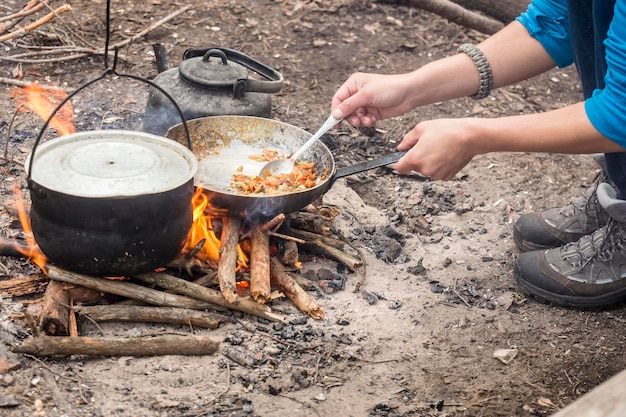 Tourist man fry food in pan over campfire in forest during hike\
cauldron hanging beside