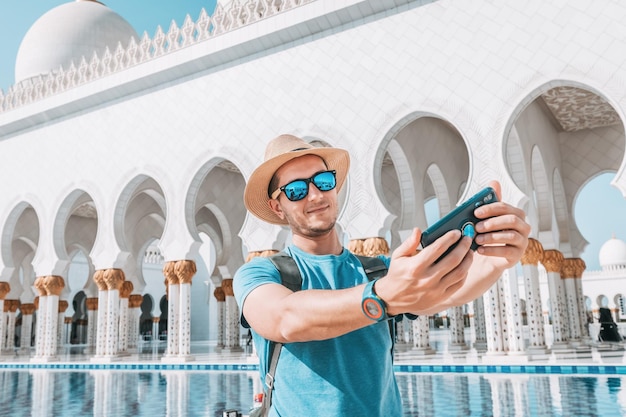 A tourist man in hat with backpack using mobile phone to make selfie photos in Great Mosque of Sheikh Zayed in Abu Dhabi