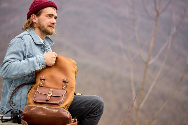 Tourist man tourist man opening leather brown backpack during hike in mountains, sit having rest, alone, side view portrait of handsome hipster guy in casual wear and hat. copy space