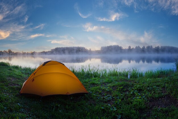 Tenda arancione turistica sull'erba verde del lago nebbioso all'alba