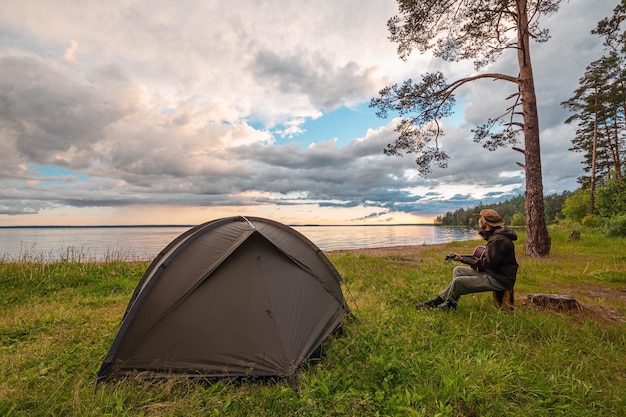 Турист играет на гитаре возле палатки на берегу озера, наслаждаясь видом. Единение с природой.