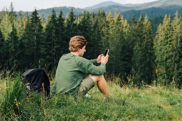 Tourist sits on the grass of a mountain meadow with a backpack and uses the Internet on a smartphone with a serious face on a background of beautiful views