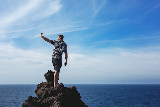 Tourist standing on a rock on background blue sea and taking photo by his smart phone camera. Lanzarote island, Spaine.