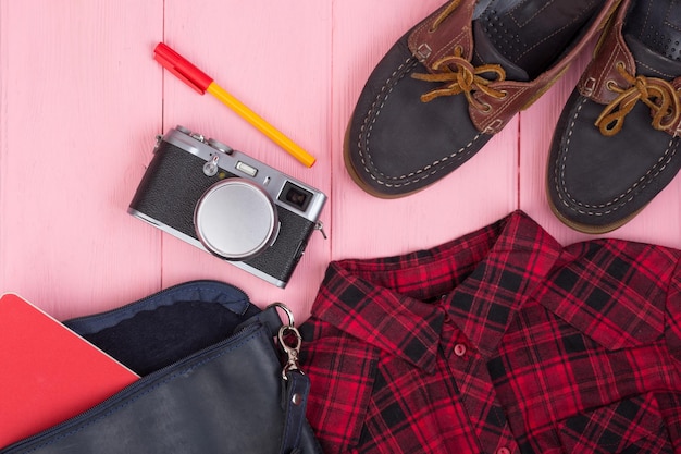 Туристическая сумка для фотоаппарата, обувь, рубашка, блокнот на розовом деревянном фоне