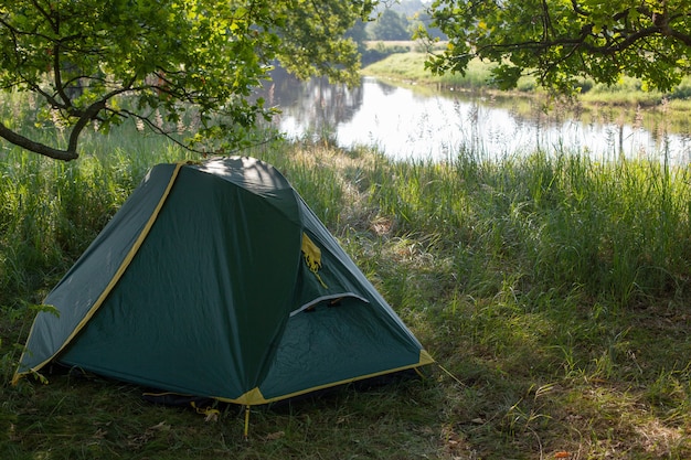 A tourist tent stands on the green grass near the river under a huge oak tree. travel wild in nature