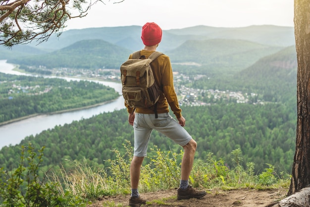 A tourist traveler with a backpack and a red hat is standing on the edge of a cliff and is looking on a green valley with the river