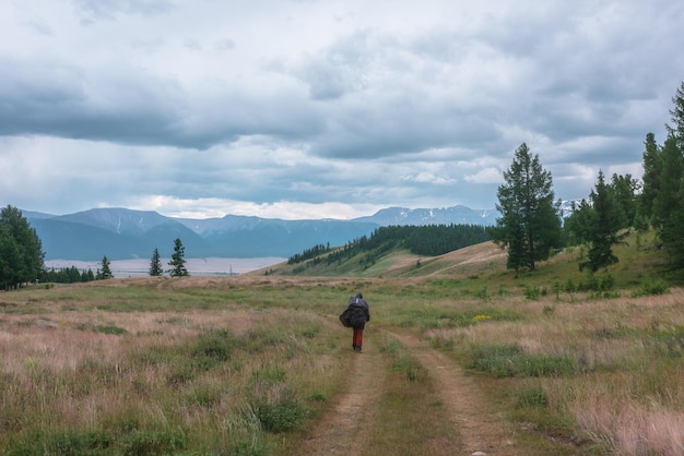 Tourist walks through hills and forest towards bad weather Hiker on way to large snow mountain range under rainy cloudy sky Man in raincoat in mountains in overcast Traveler goes towards adventure