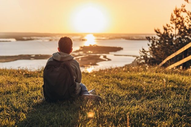 Tourist with backpack sitting on top of hill in grass field and enjoying beautiful landscape view Rear view of teenage boy hiker resting in nature Active lifestyle Concept of local travel