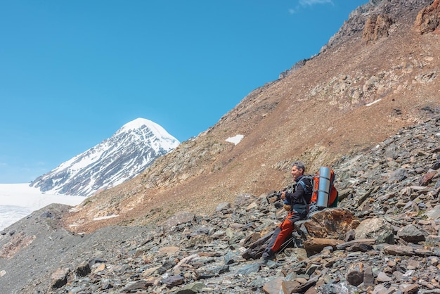 Turista con macchina fotografica seduto su pietra in alta montagna in una giornata di sole scenico paesaggio alpino con l'uomo sotto il sole splendente contro la vetta della montagna innevata e le rocce alla luce del sole zaino in spalla tra le montagne innevate