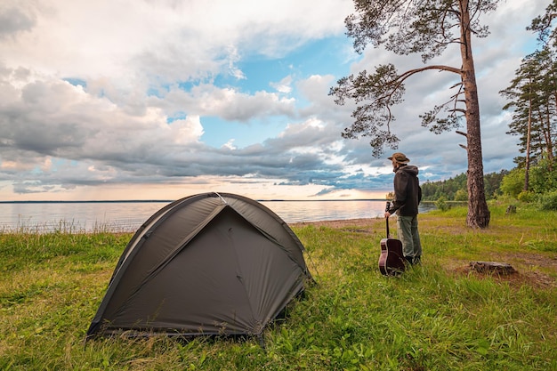 Tourist with guitar standing near camp tent on lake shore enjoying the view at sunset