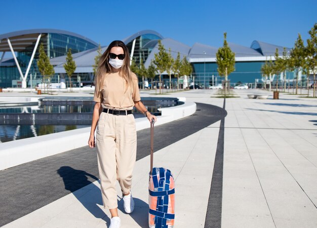 Tourist woman in madical mask walking with luggage near airport building