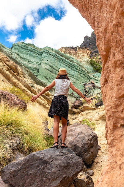 A tourist woman with a hat and open arms at the Azulejos de Veneguera or Rainbow Rocks in Mogan Gran Canaria