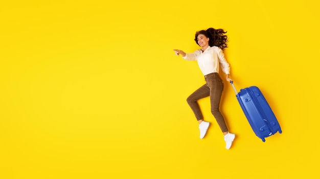 Tourist woman with travel suitcase pointing aside on yellow background