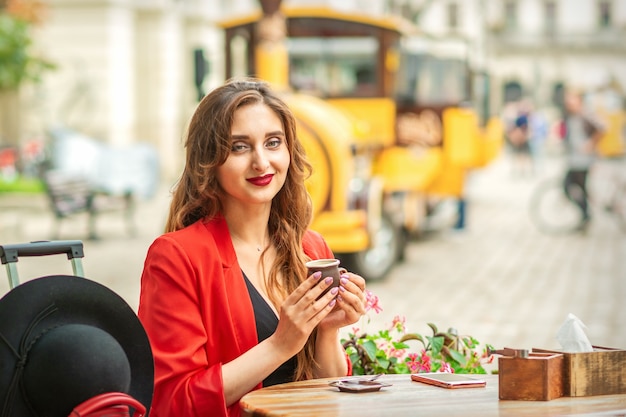 Tourist young caucasian woman in red jacket with coffee cup at the table in cafe outdoors