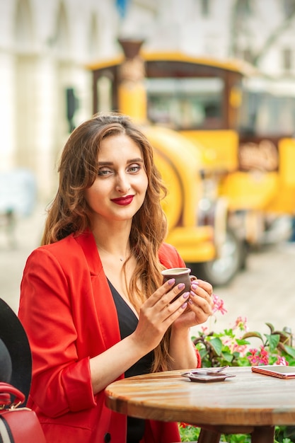 Tourist young caucasian woman in red jacket with coffee cup at the table in cafe outdoors.