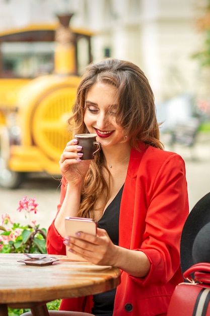 Tourist young caucasian woman in red jacket with coffee cup at the table in cafe outdoors.