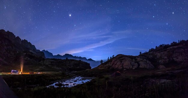 Tourists and hunters are heated around campfire at night in mountains in background of starry sky