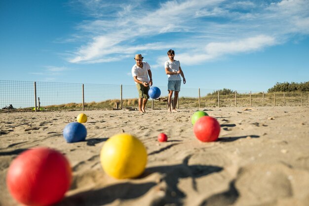 Photo tourists play an active game petanque on a sandy beach by the sea - group of young people playing boule outdoors in beach holidays - balls on the ground