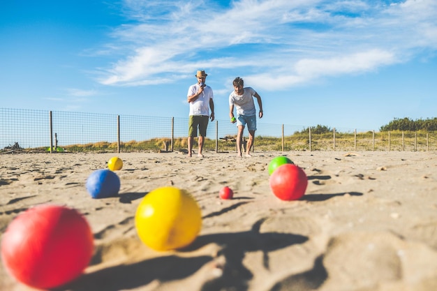 Photo tourists play an active game petanque on a sandy beach by the sea - group of young people playing boule outdoors in beach holidays - balls on the ground