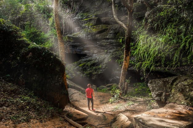 Tourists stand in front of a large rock with beams of light shining down on the tropical forest at Tat Fa Waterfall, the best waterfall within Phu Wiang National Park, Khon Kaen, Thailand.