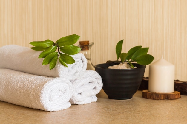 Towels, cosmetic massage oil, leaf, sea salt with shells in black bowl and candle on a wooden stand.