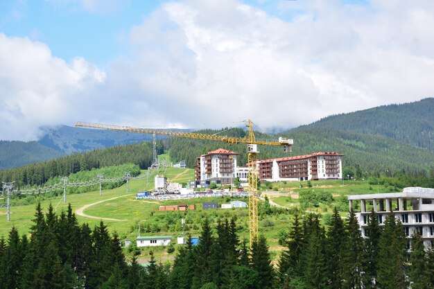 A tower crane builds a ski resort in the mountains in summer, against the backdrop of mountains, forests and hotels under construction. The ski lifts and the blue sky are visible