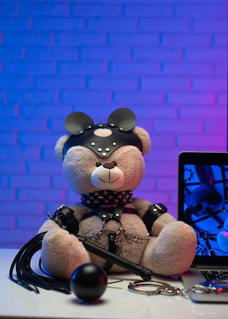 The toy bear in a leather belt accessory for bdsm games next to a laptop tv in neon colors