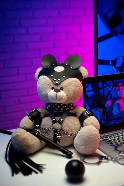 The toy bear in a leather belt accessory for BDSM games next to a laptop TV in neon colors