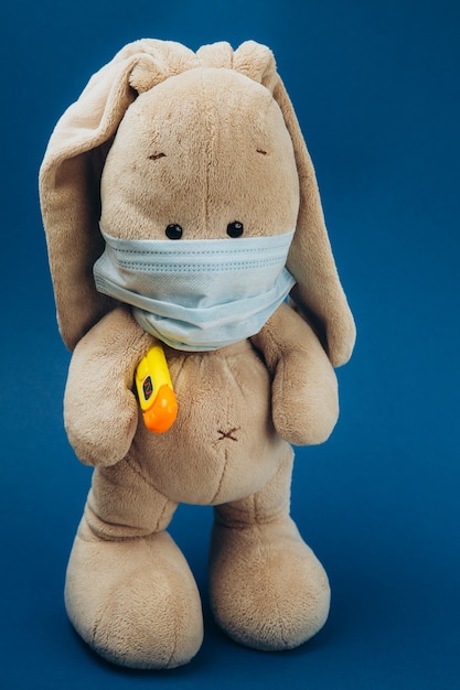 Toy bunny with thermometer under paw and in mask on a blue surface