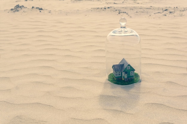 Toy cardboard house with green lawn protected by a glass dome cloche among of a lifeless sand desert