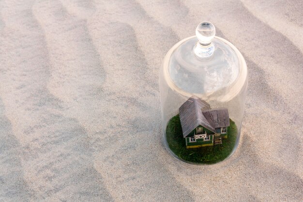 Toy cardboard house with green lawn protected by a glass dome cloche among of a lifeless sand desert