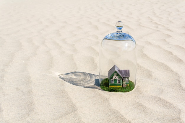活気のない砂の砂漠の中でガラスのドームクローシュで保護された緑の芝生のおもちゃの段ボールの家