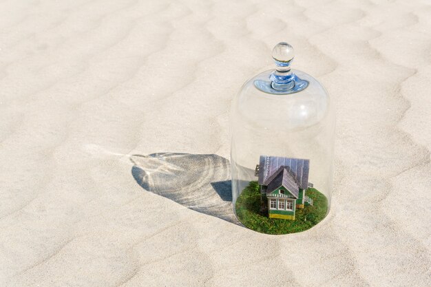 Toy cardboard house with green lawn protected by a glass dome cloche among of a lifeless sand desert