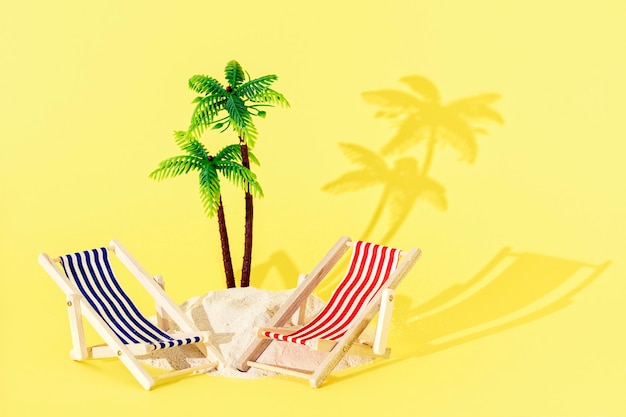 Toy mini beach deck chairs and palm tree with sand and shadow on yellow background. Creative summer beach concept.