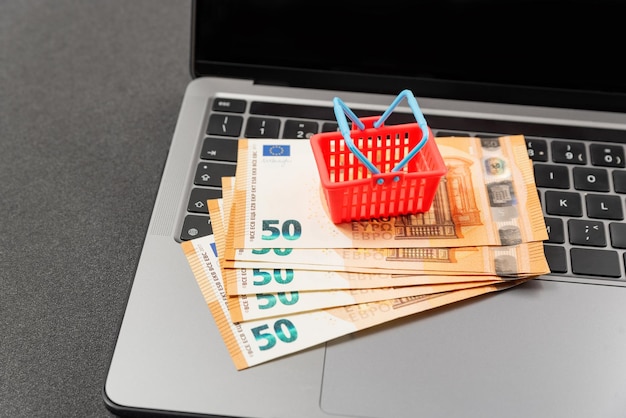 Toy miniature shopping cart and cash euros on laptop keyboard Concept of online shopping Cost of grocery basket in Europe