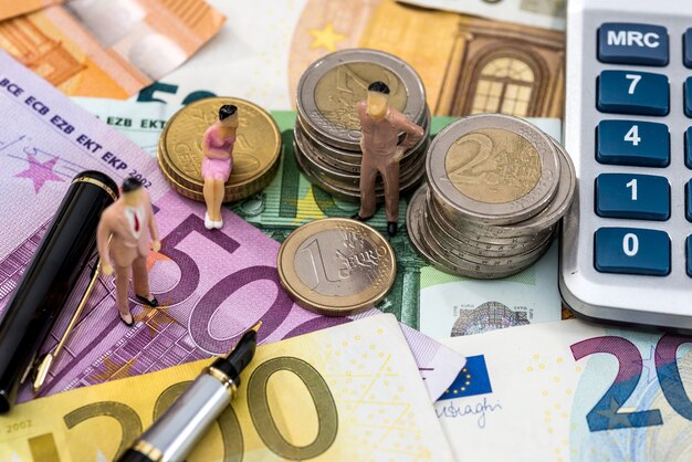 Toy people sit on euro coin with calculator, pen and euro bills