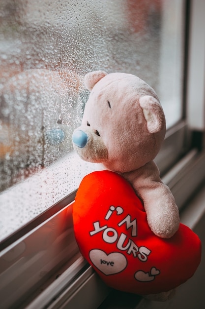 A toy pink sad bear with a red plush heart sitting on the windowsill Autumn rainy day Raindrops