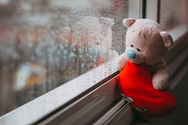 A toy pink sad bear with a red plush heart sitting on the windowsill Autumn rainy day Raindrops