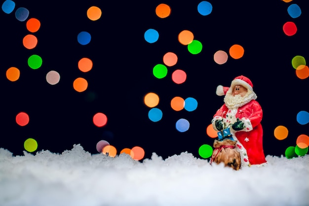 Toy Santa Claus in red coat with bag of gifts by night in the background of colorful bokeh lights